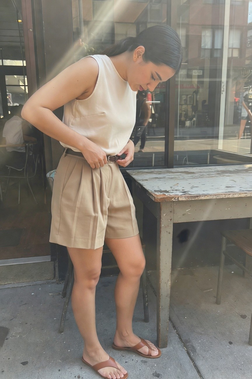 Woman adjusting brown belt on beige pleated shorts with white sleeveless tank top and brown flip-flops, standing outdoors near a table