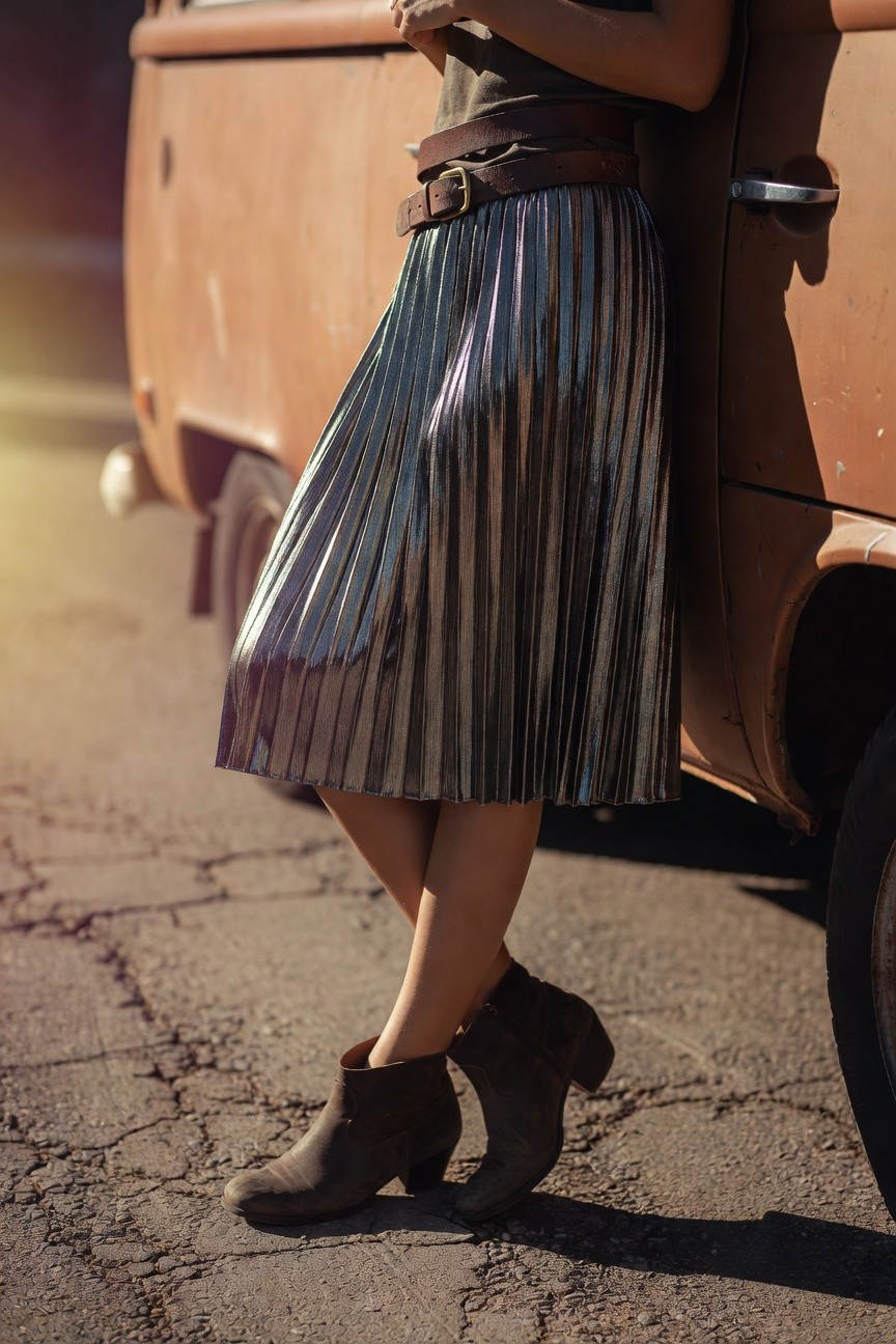 Woman posing next to a vintage van in an olive green cropped top, brown leather belt with gold buckle, silver metallic pleated midi skirt, and brown suede ankle booties on a cracked pavement surface