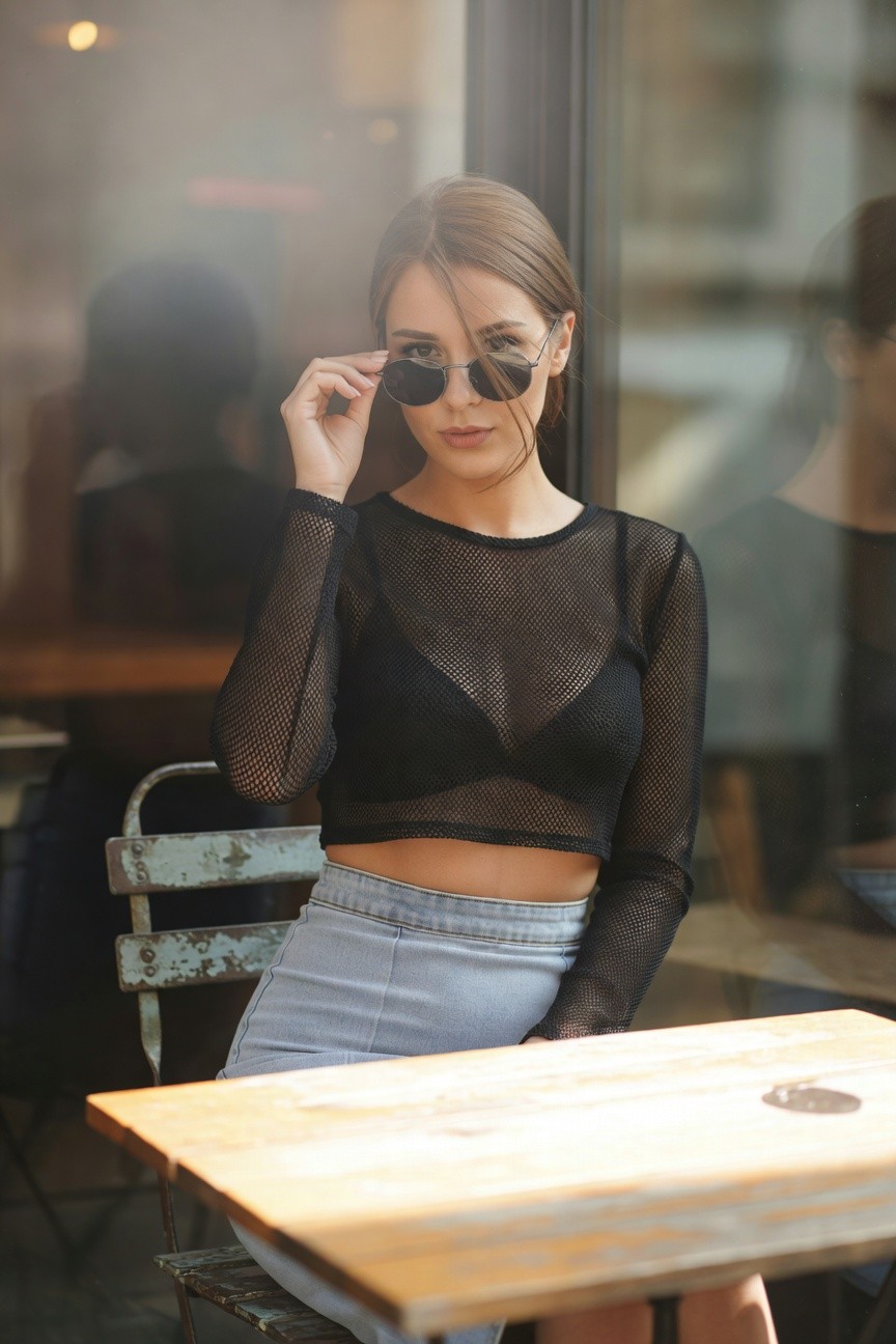 Woman wearing sheer black long-sleeve mesh crop top and light wash high-waisted denim mini skirt, hand adjusting round sunglasses on nose, seated pose