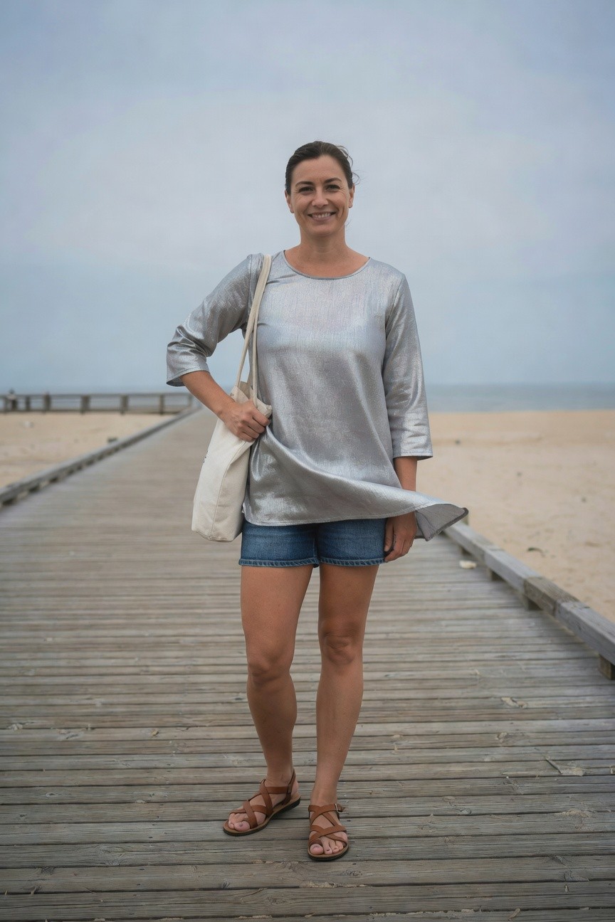 Woman standing on wooden pier in shiny silver metallic tunic dress over short blue denim cutoffs, holding white canvas tote bag, wearing tan gladiator strap sandals, smiling at camera