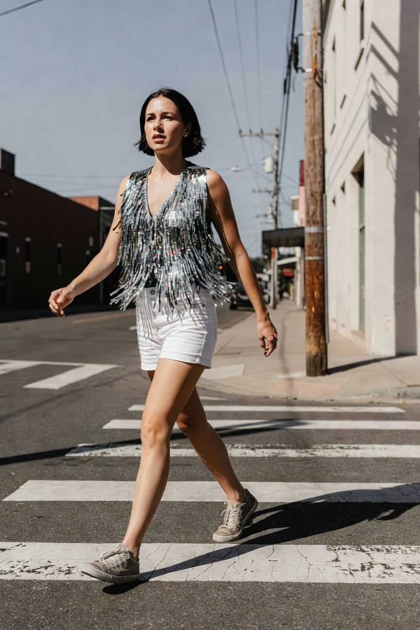 Woman crossing street in silver sequin fringed tank top, white denim shorts, and tan sneakers, urban daytime setting