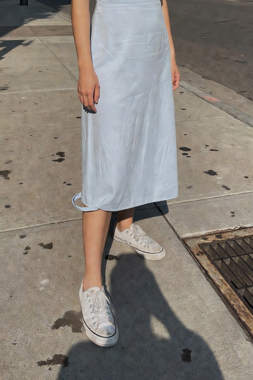 Woman in a light blue flowy midi dress standing on a sidewalk, paired with white Converse sneakers, side view focusing on the skirt's drape and casual footwear
