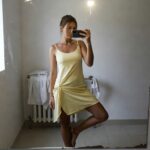 Woman taking mirror selfie in bathroom wearing pale yellow spaghetti-strap mini sundress with hand near hem, one leg extended wearing metallic strappy flat sandals, beige towel draped on sink nearby
