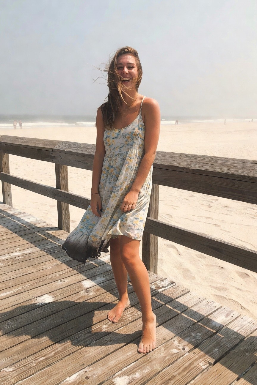 Smiling woman with long wavy brown hair wearing a white floral spaghetti strap sundress with ombre skirt hem, black bag, thin bracelet and necklace, standing barefoot on wooden boardwalk railing