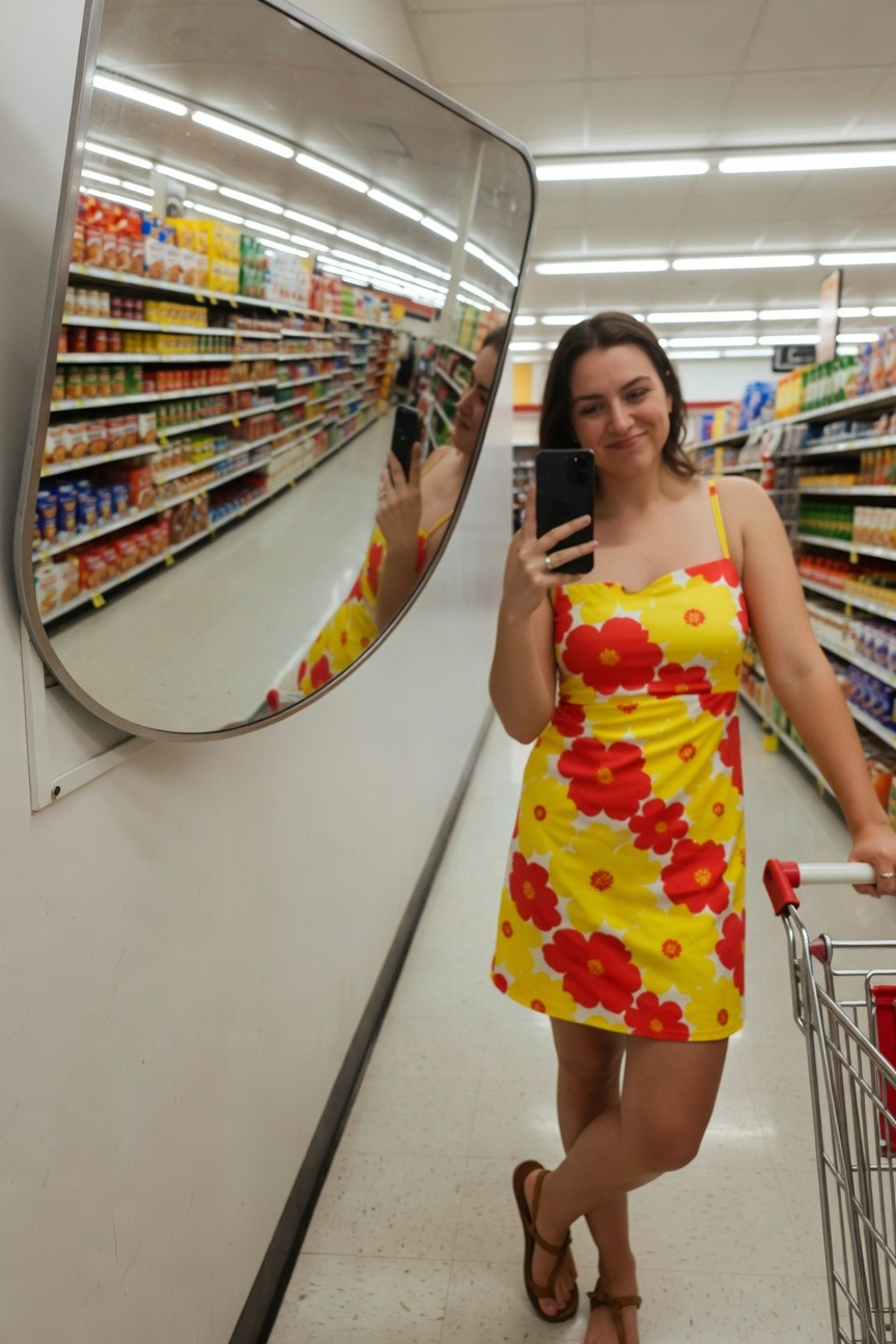 Woman smiling in mirror selfie wearing yellow sundress with large red flowers, spaghetti straps, holding red-handled shopping cart in grocery store aisle, tan strappy sandals on feet