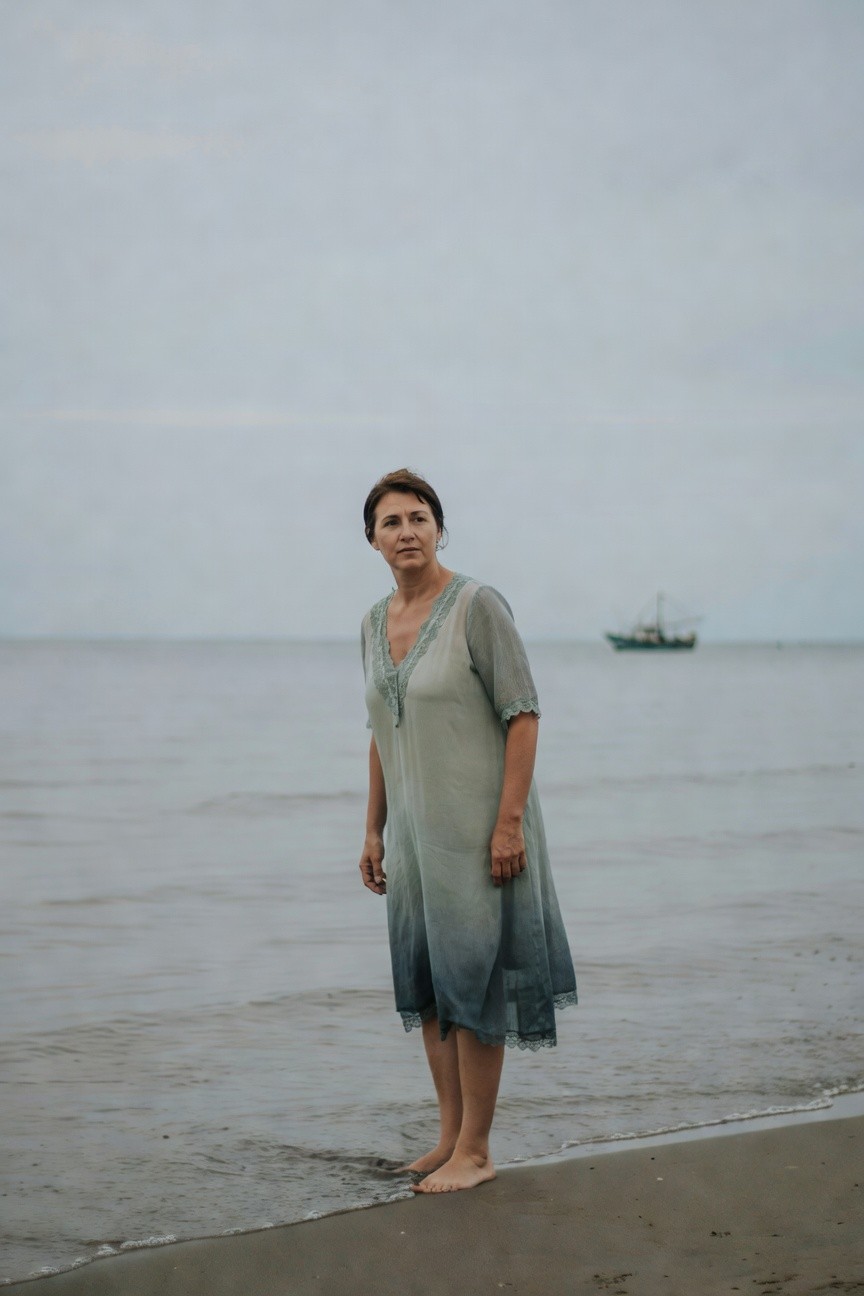 Woman in a sheer gradient chiffon maxi dress fading from light gray to teal with lace trim at V-neck, short sleeves, and scalloped hem, standing barefoot on a beach with ocean and boat in blurred background