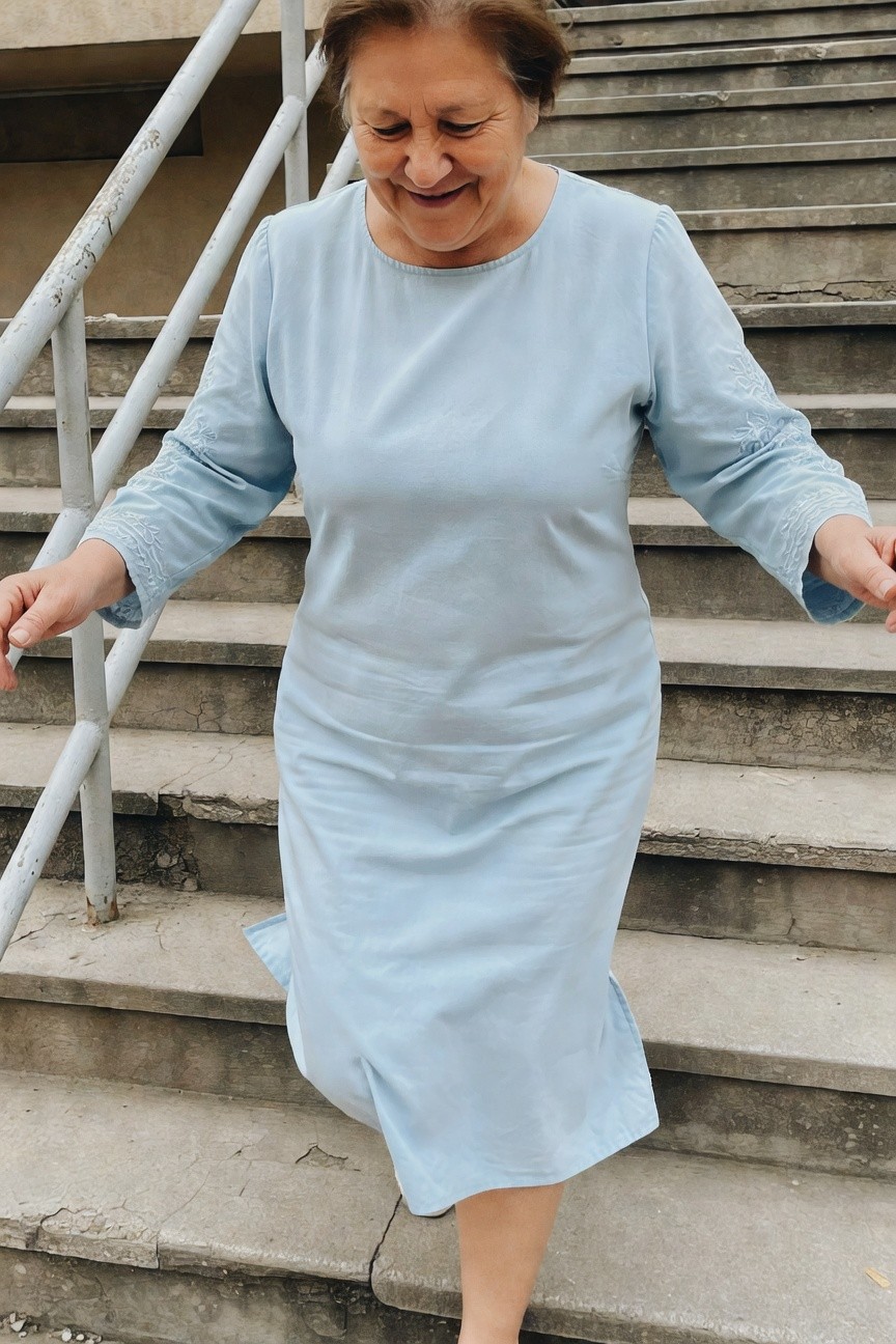 Smiling older woman in a light blue long-sleeved midi dress with white floral embroidery on cuffs, ascending concrete outdoor stairs while holding metal handrails