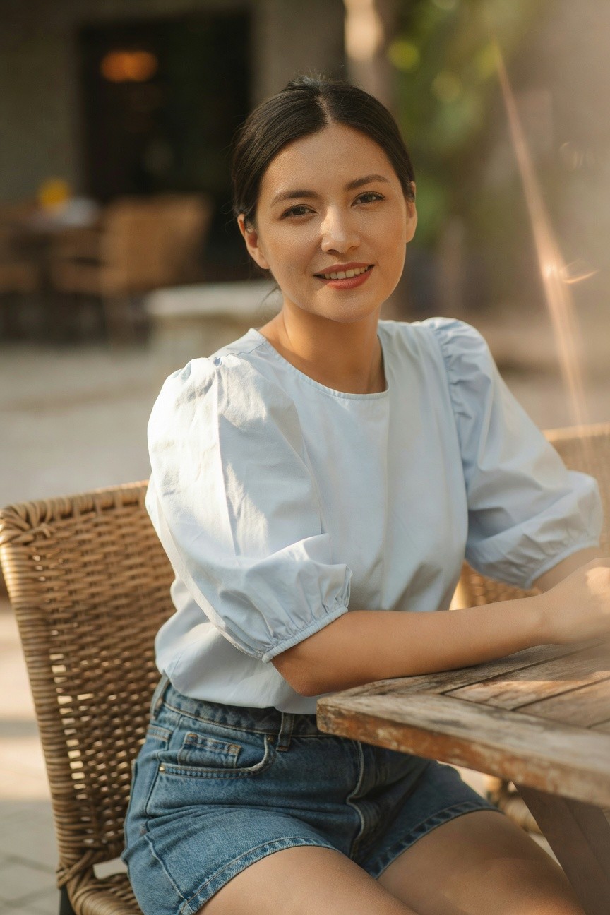 Woman in light blue puffed sleeve blouse and high-waisted denim shorts, seated at a wooden table with arms resting on it