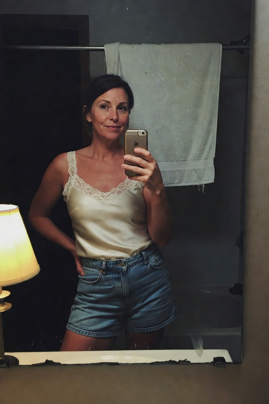 Woman in bathroom mirror selfie wearing cream silk camisole with lace trim and light wash high-waisted denim shorts, hands on hips smiling at camera