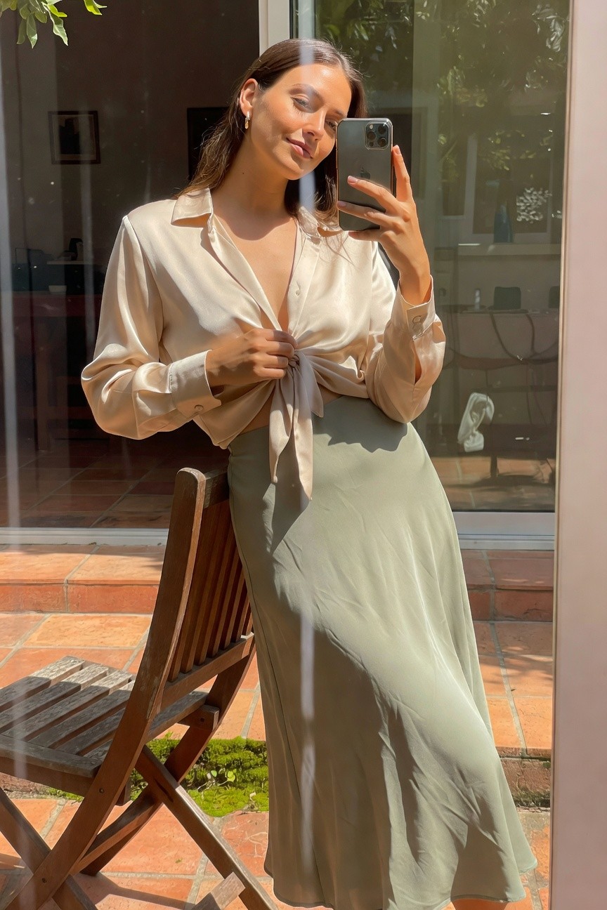 Woman in selfie wearing open cream silk long-sleeve blouse tied at waist over sage green flowy midi skirt, gold hoop earrings, leaning against wooden chair on terrace