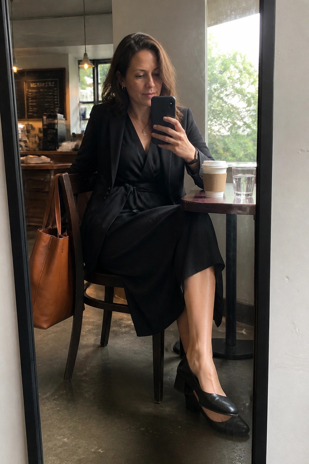 Woman in black blazer and dress taking mirror selfie in cafe with coffee and bag.