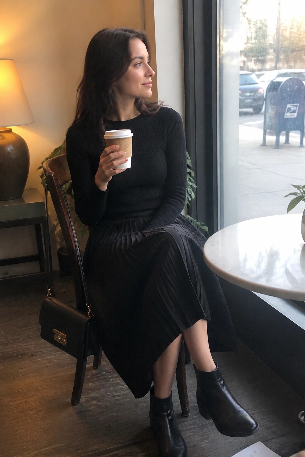 Woman in black sweater and pleated skirt holding coffee by cafe window
