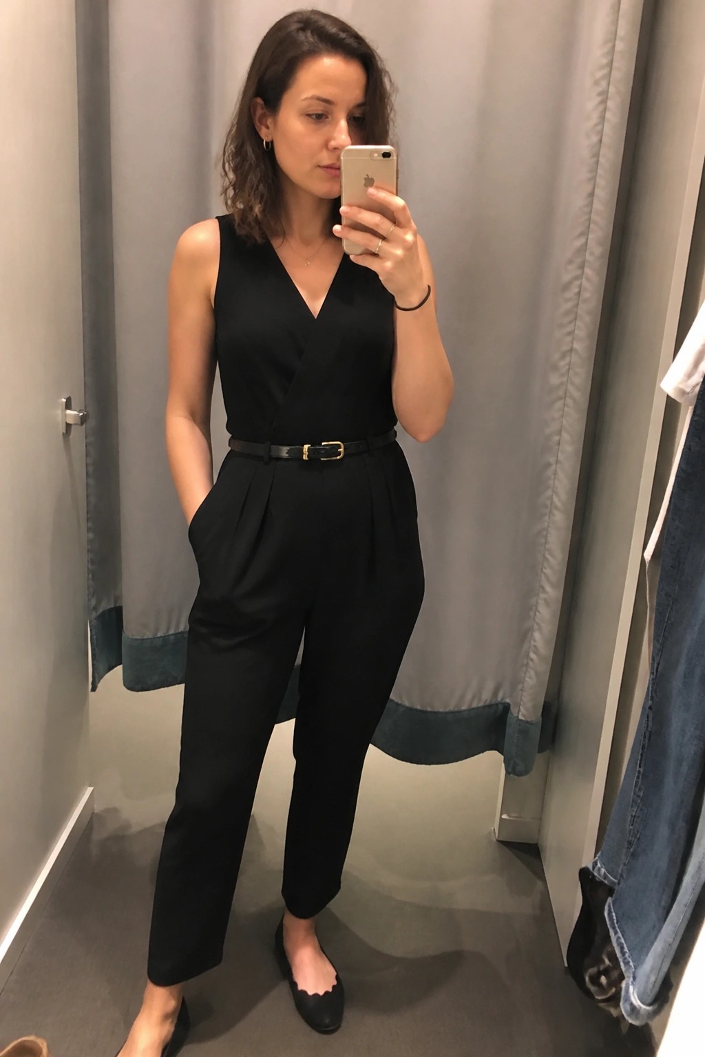 Woman in black sleeveless jumpsuit with thin belt and black flats taking a mirror selfie in a fitting room.