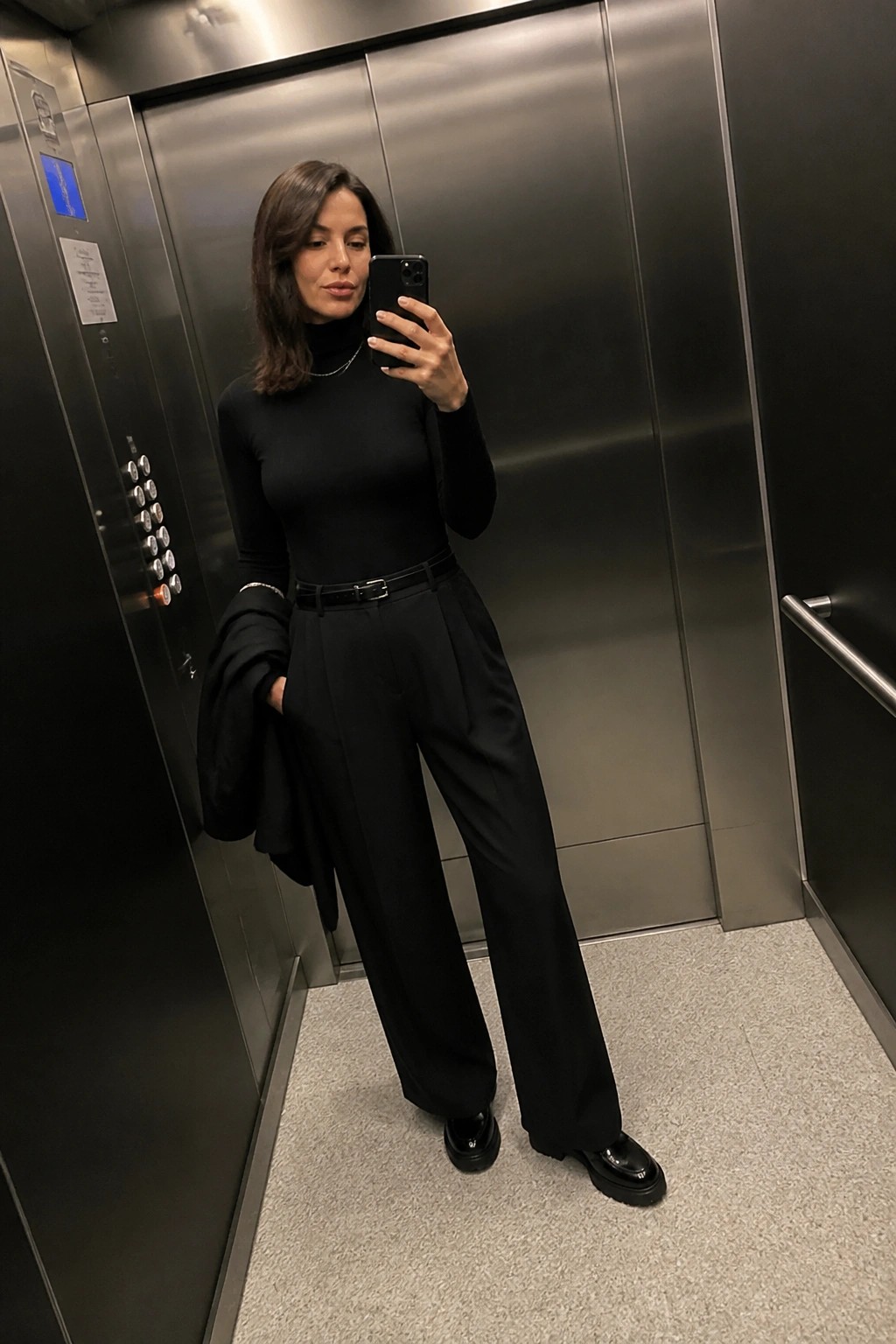 Woman in black turtleneck and pleated trousers taking selfie in elevator