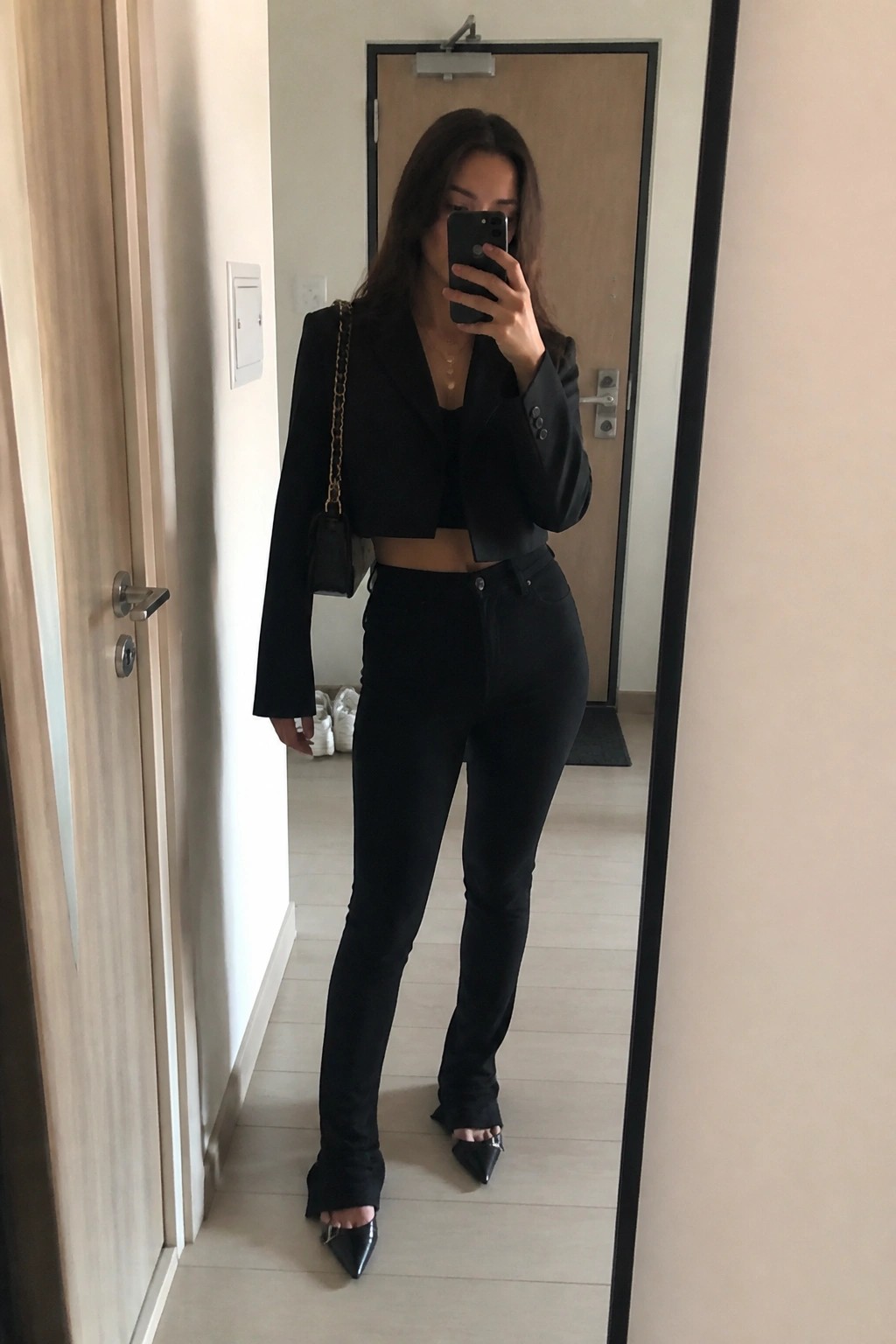 Woman in all black outfit with cropped blazer over crop top, slim black jeans, pointed toe heels with chain detail, and small black handbag with gold chain strap, taking a mirror selfie.