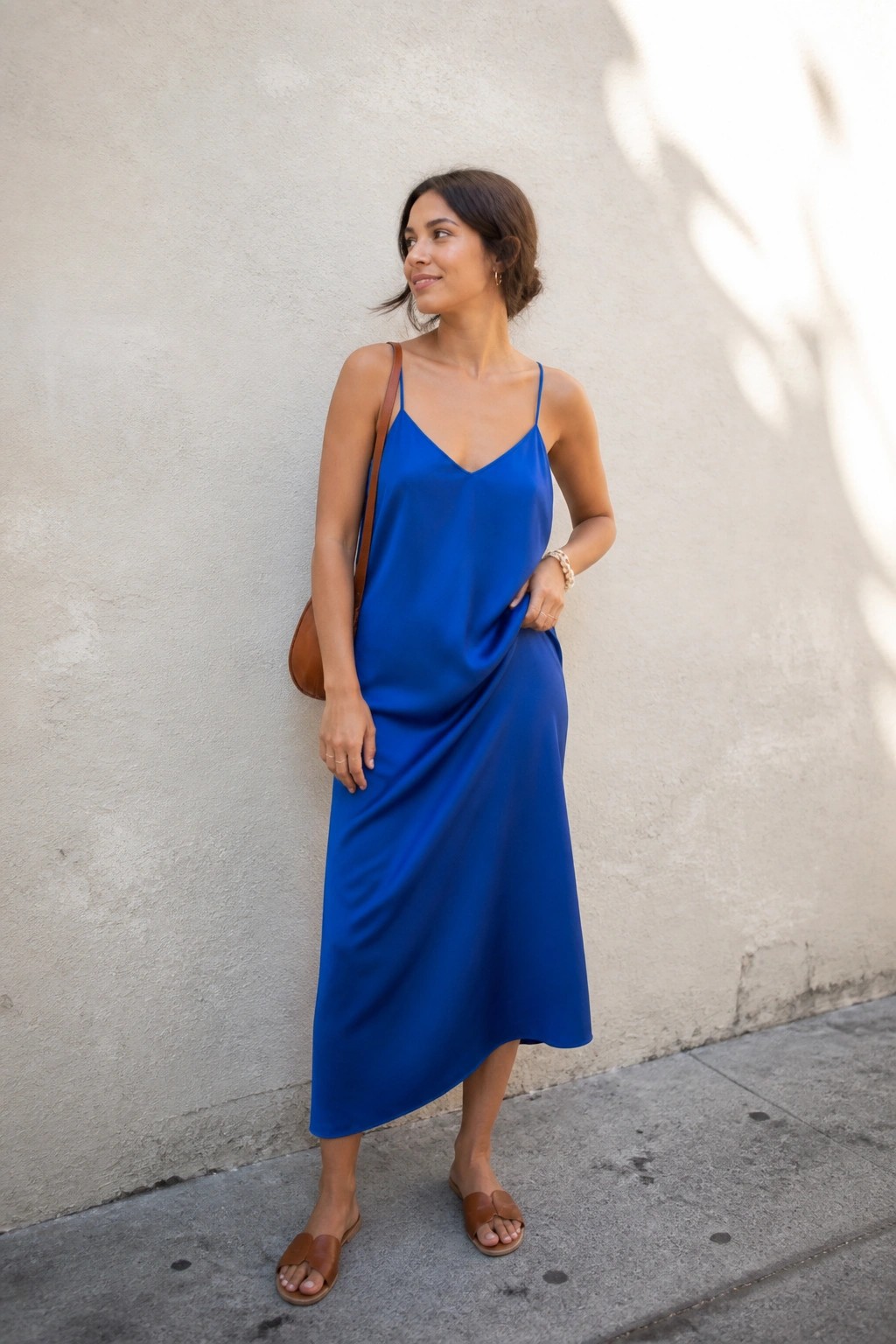 Woman in blue satin slip dress with brown bag leaning against wall on sidewalk