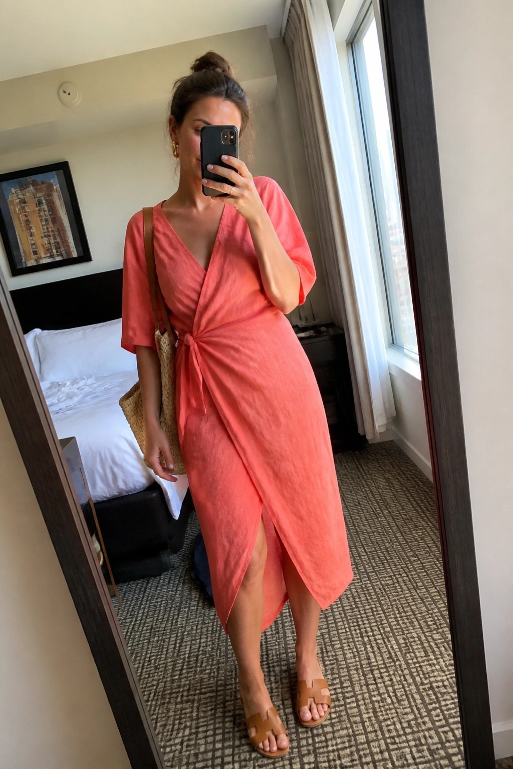 Woman wearing a coral linen wrap midi dress with short sleeves and side knot tie, paired with brown leather flat sandals and a woven straw tote bag, shown in a mirror selfie inside a hotel room.