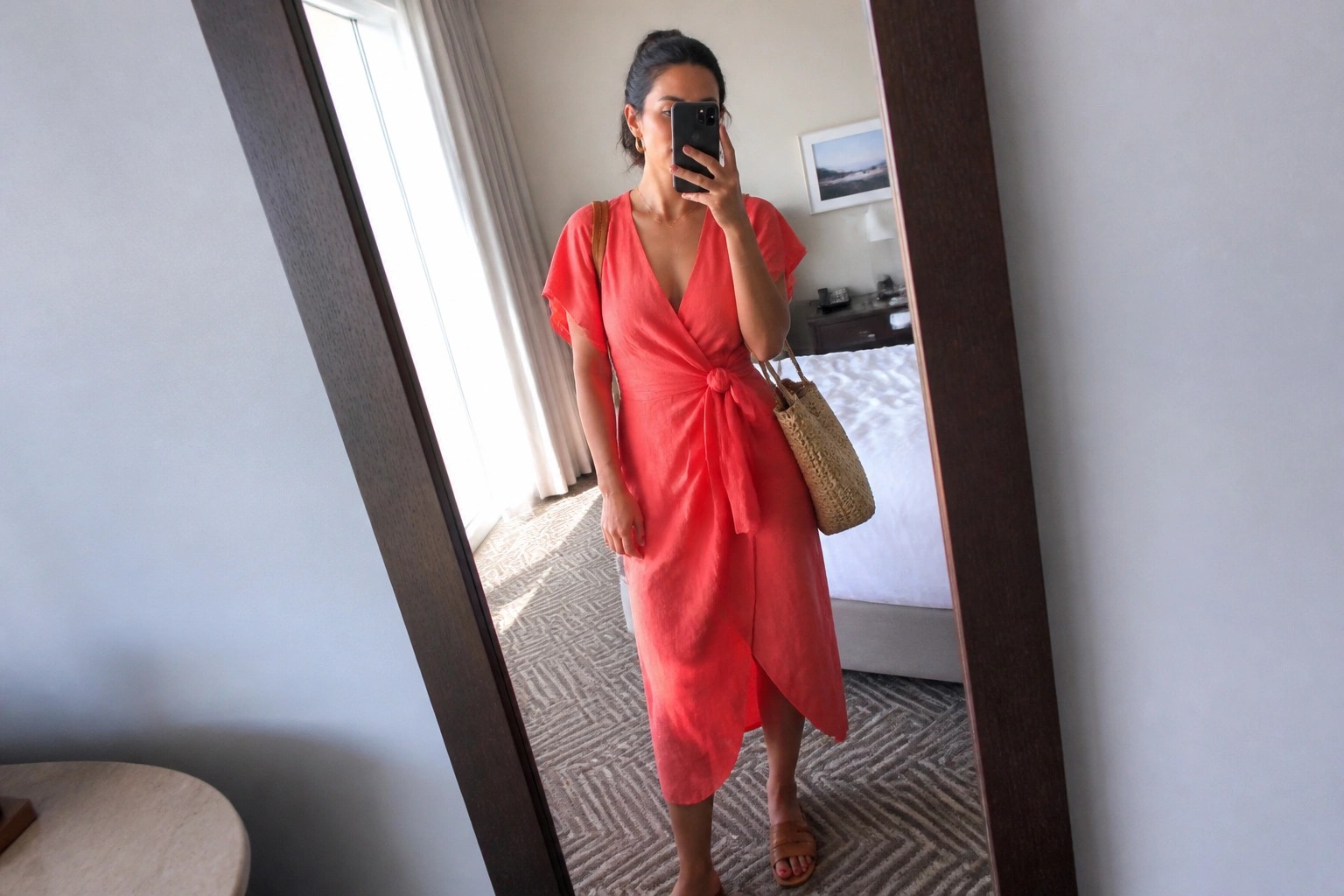 Woman wearing a coral linen wrap midi dress with short sleeves and side knot tie, paired with brown leather flat sandals and a woven straw tote bag, shown in a mirror selfie inside a hotel room.