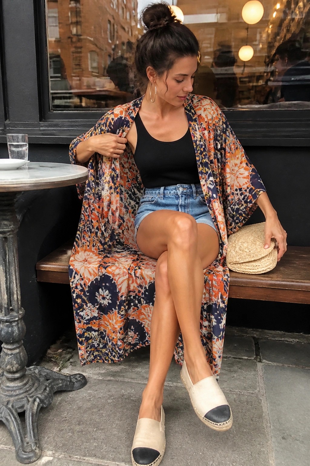 Woman in floral kimono, black tank, and denim shorts sits with straw clutch outdoors