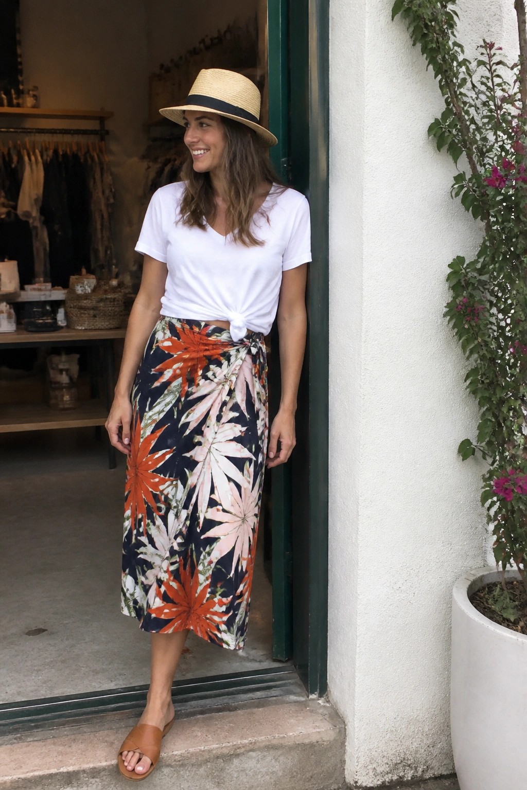 Woman in straw hat, knotted white tee, and orange floral wrap skirt in doorway