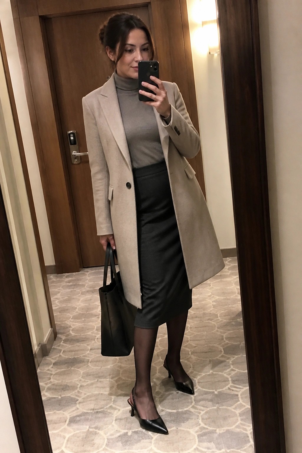 Woman in beige coat over gray turtleneck and black skirt taking mirror selfie with tote bag.