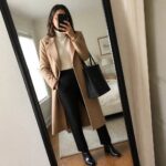Woman in tan coat, cream sweater, black pants holding tote in mirror selfie.