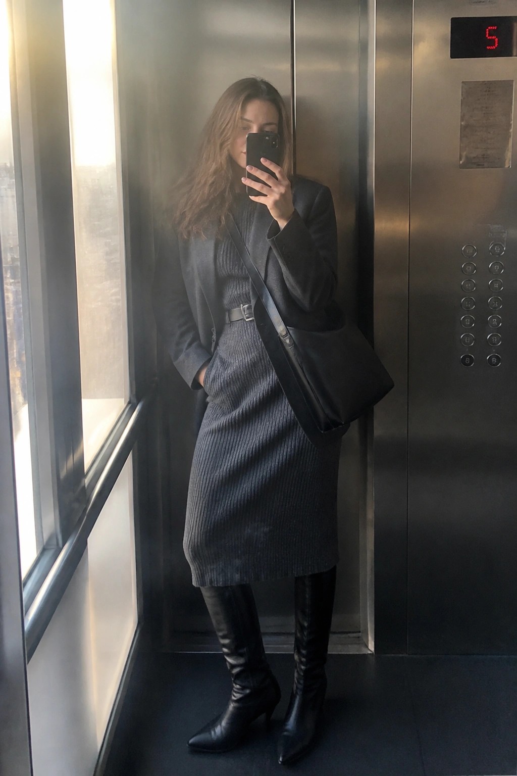 Woman in gray blazer and ribbed dress taking selfie in elevator with black boots and bag