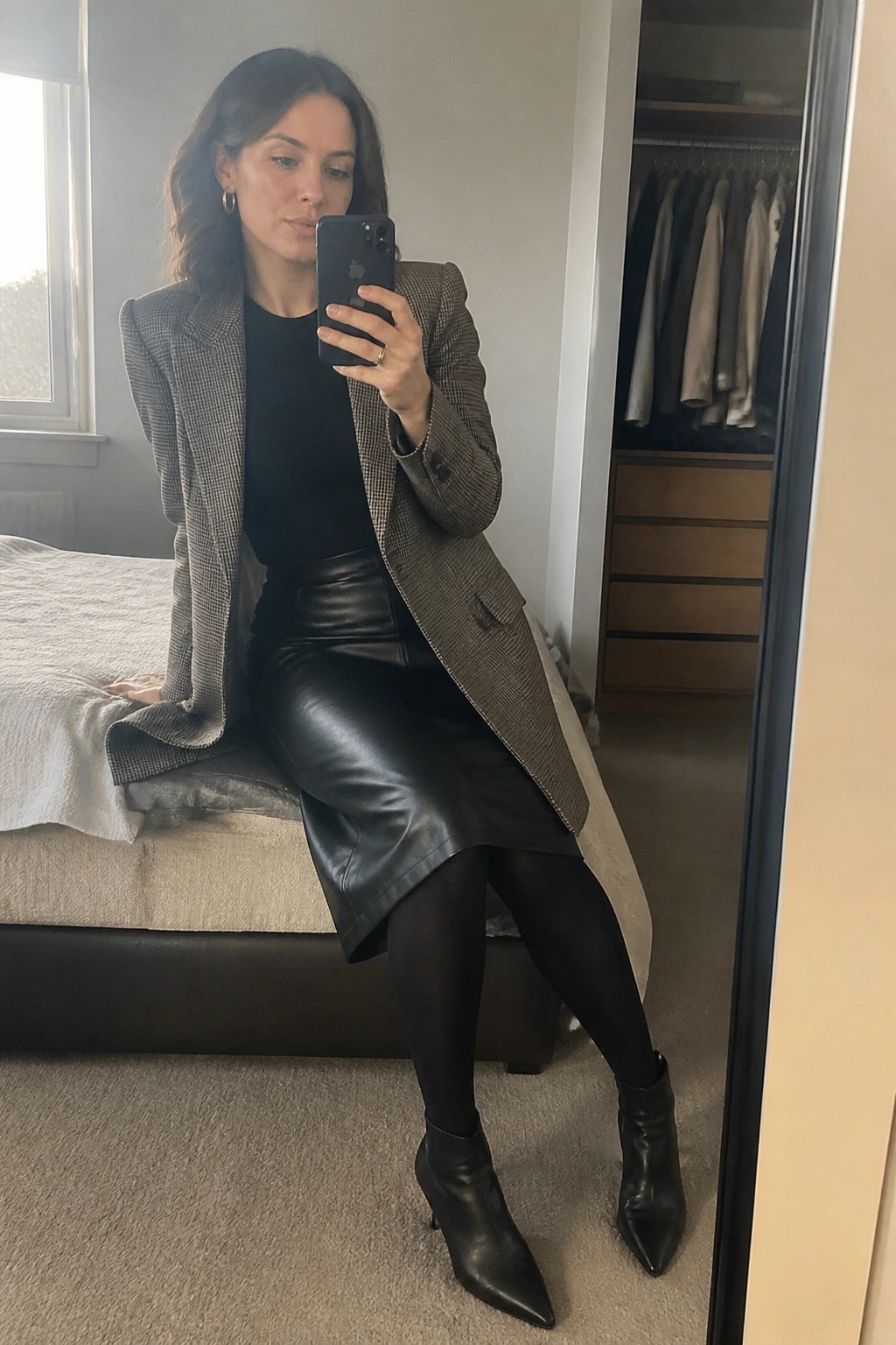 Woman in checkered blazer and leather skirt taking bedroom mirror selfie with phone