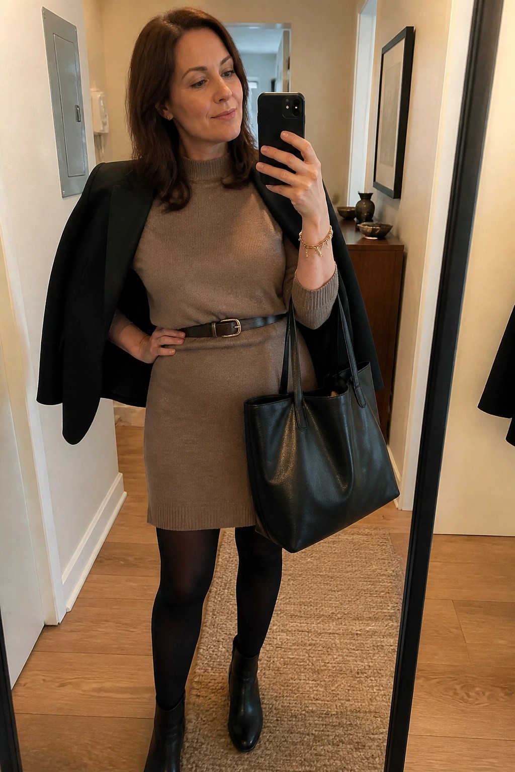 Woman in taupe sweater dress and black blazer taking mirror selfie with tote bag