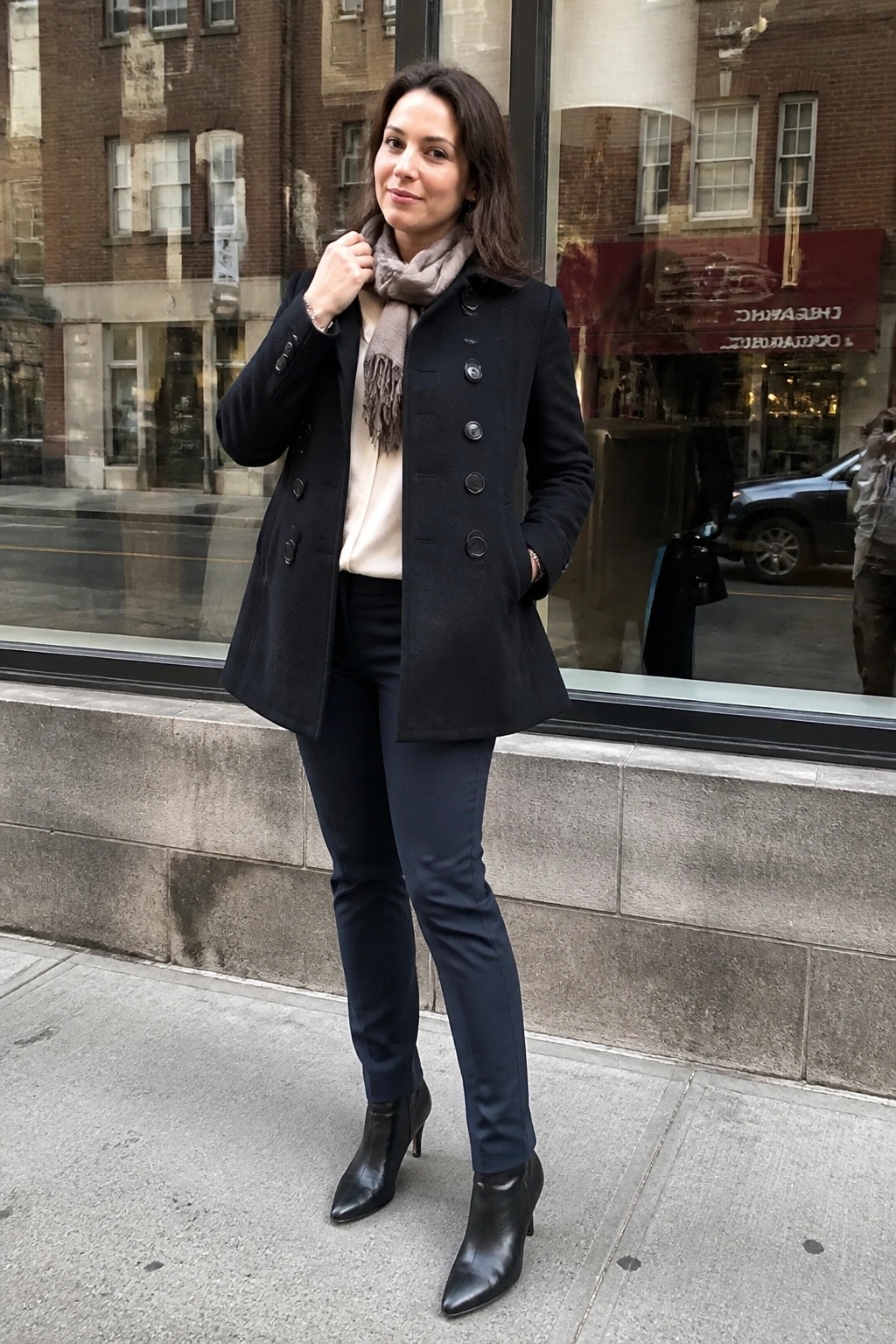 Woman in black coat and gray scarf stands on city sidewalk in boots
