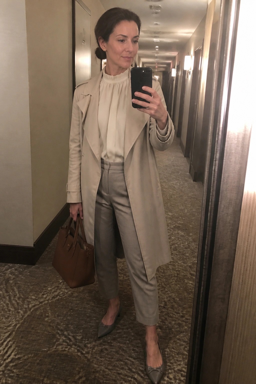 Woman in beige trench coat and gray pants taking mirror selfie with brown bag.