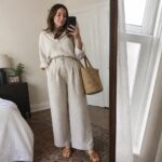 Woman in cream linen shirt and pants holding phone and woven bag in bedroom