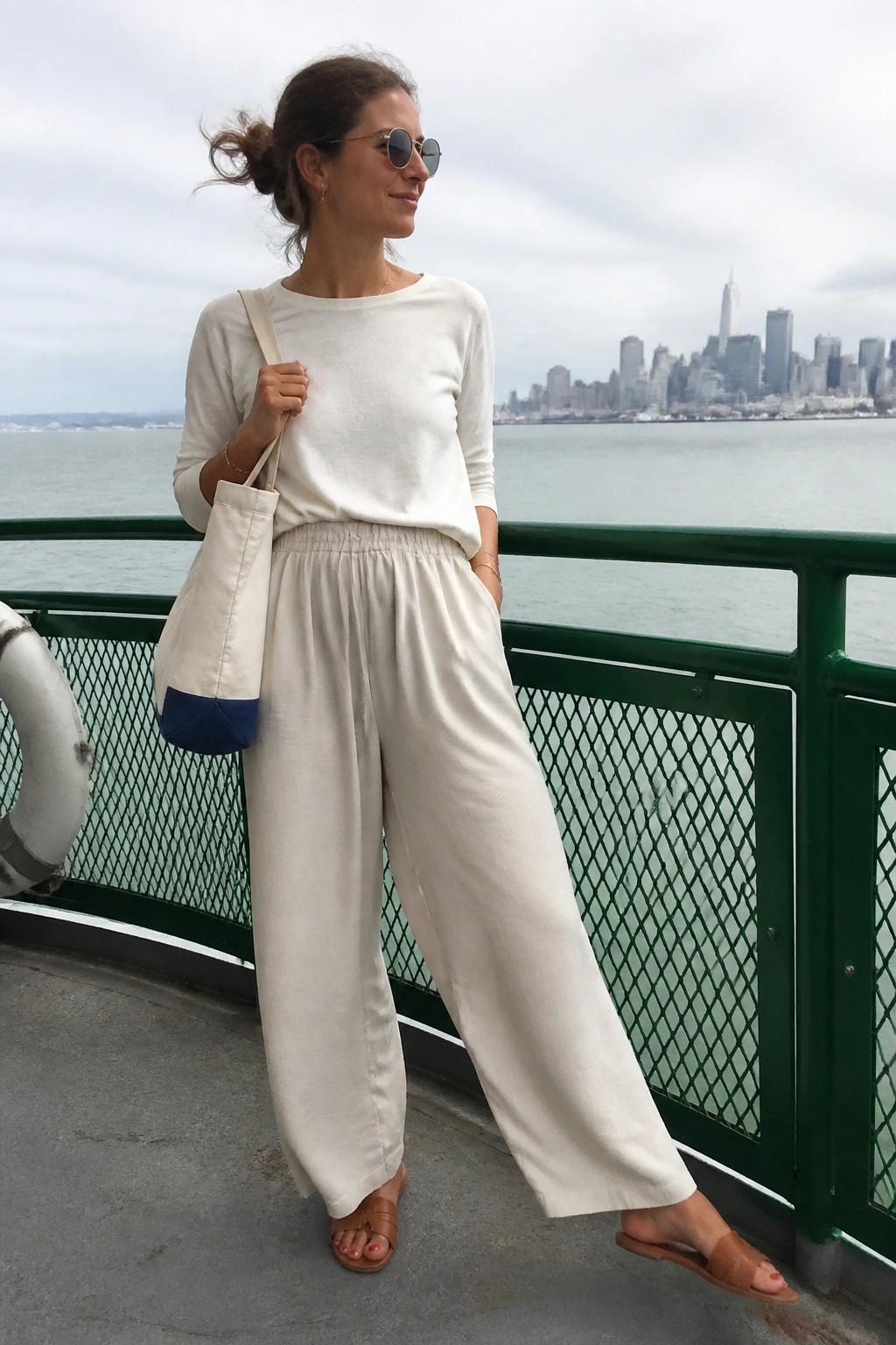 Woman in an all-cream outfit featuring a long sleeve boat neck top tucked into wide leg elastic waist pants, brown slide sandals, and a canvas tote bag with a blue base accent, standing on a green railing with water behind her.