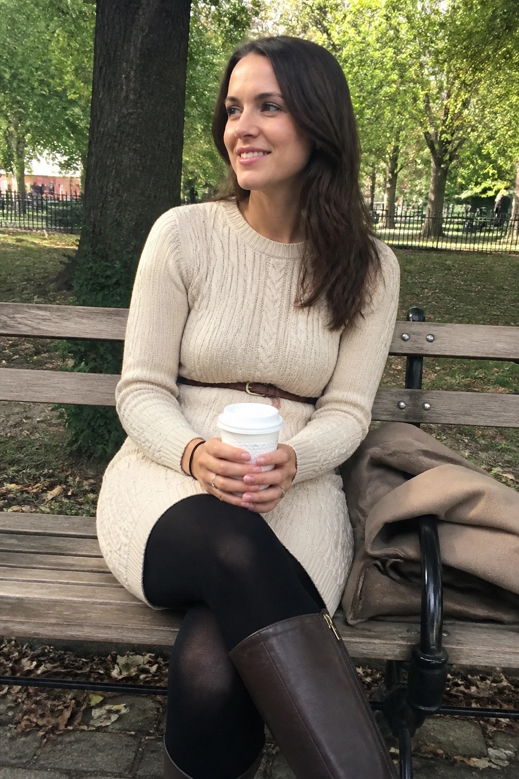Woman seated on park bench wearing cream cable knit sweater dress with brown leather belt, black tights, and brown knee high boots while holding a white coffee cup.