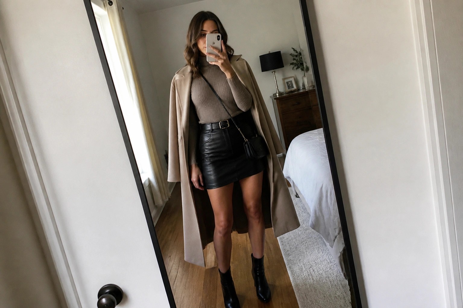 Woman in mirror selfie wearing open gray long coat over beige ribbed turtleneck sweater tucked into black leather mini skirt with black belt, black pointed ankle boots, and small black crossbody bag on gold chain.