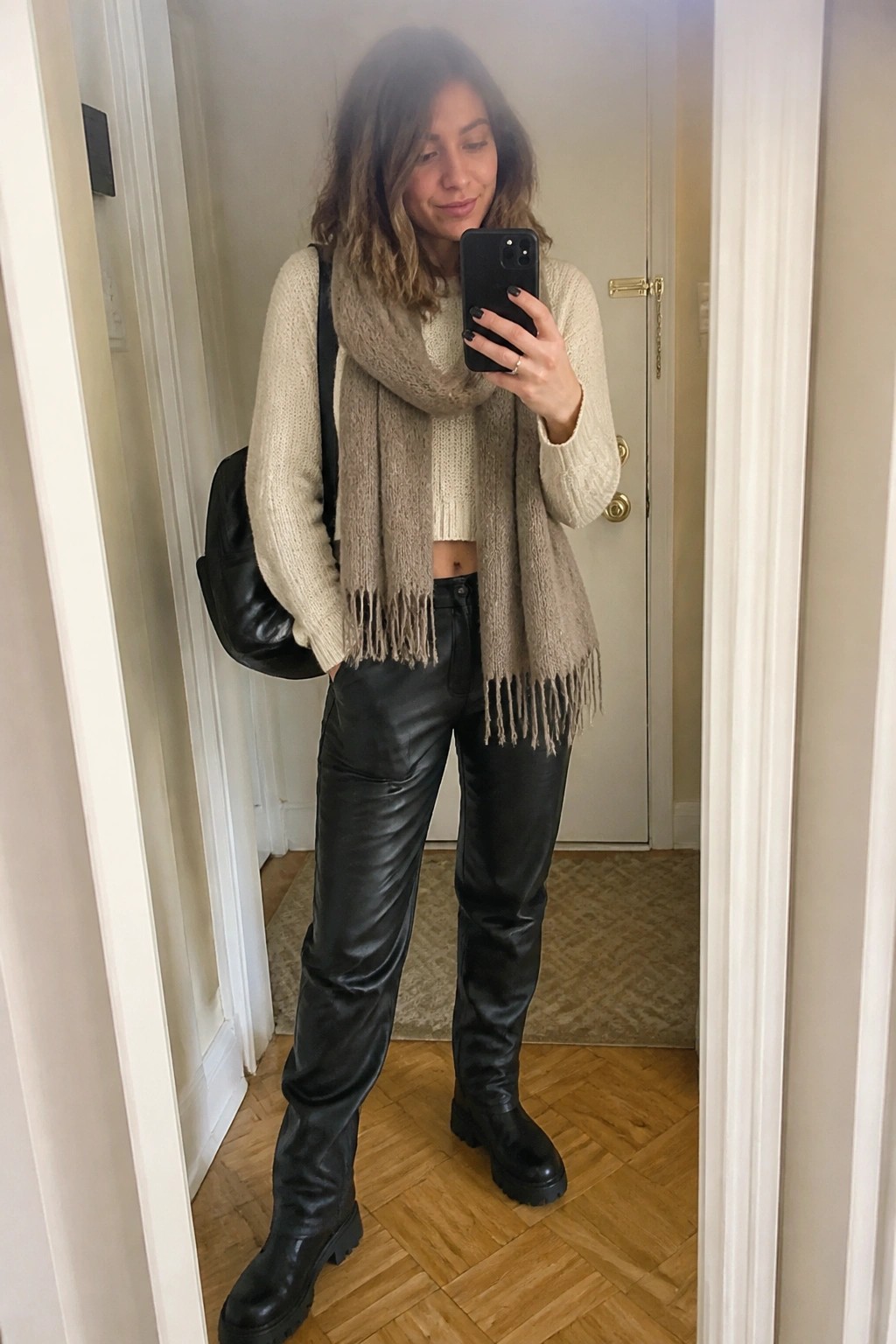 Woman taking a mirror selfie wearing a cream knit sweater, beige fringed scarf, black leather pants, and black chunky boots with a black shoulder bag.