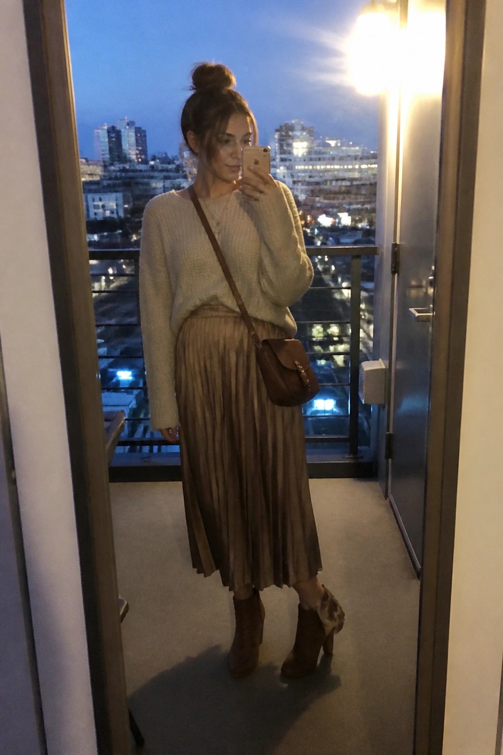 Woman in cream sweater and pleated skirt taking balcony selfie at dusk