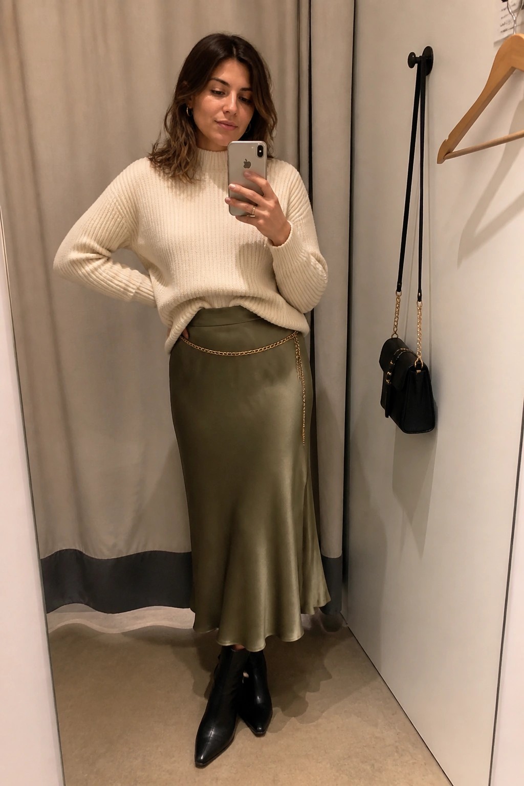 Woman in cream sweater and olive satin skirt taking mirror selfie with gold chain belt