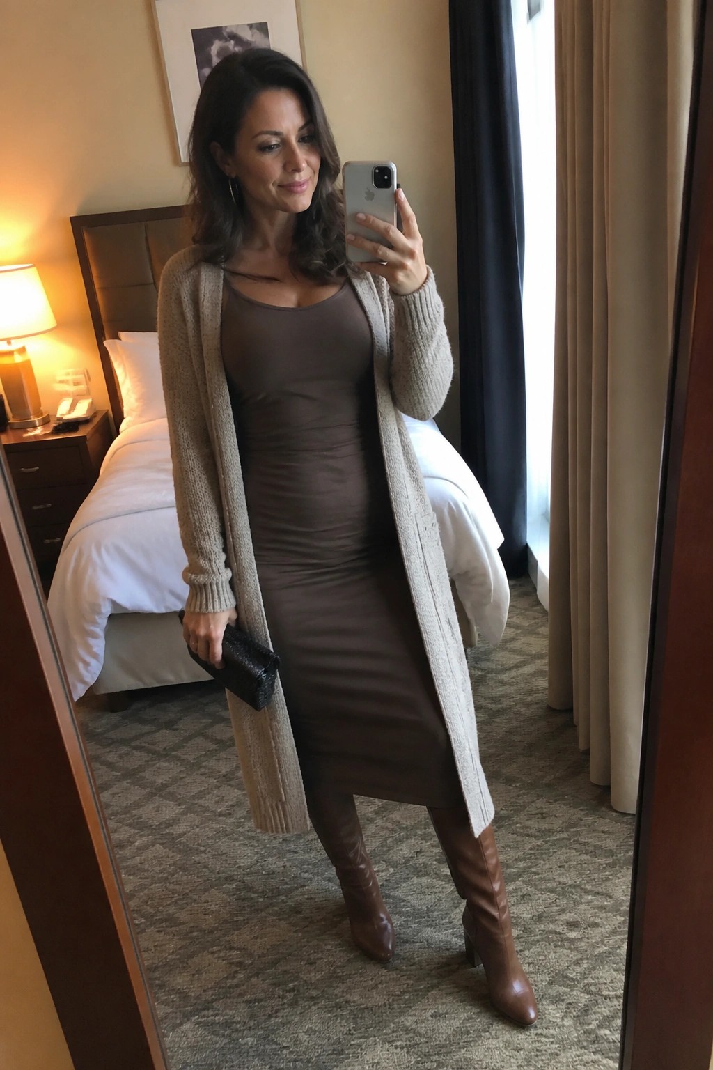 Woman in brown dress and beige cardigan taking mirror selfie in hotel room with brown boots