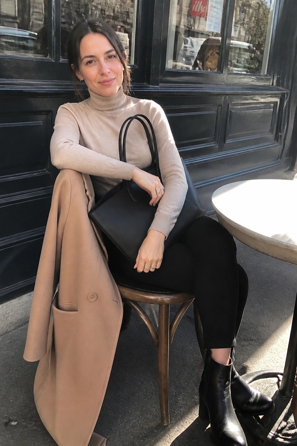 Woman in beige turtleneck holding black bag with tan coat outdoors