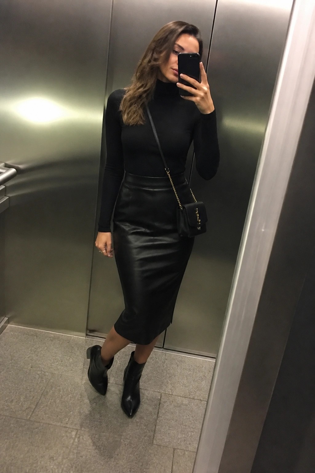Woman in black turtleneck and leather skirt taking mirror selfie in elevator