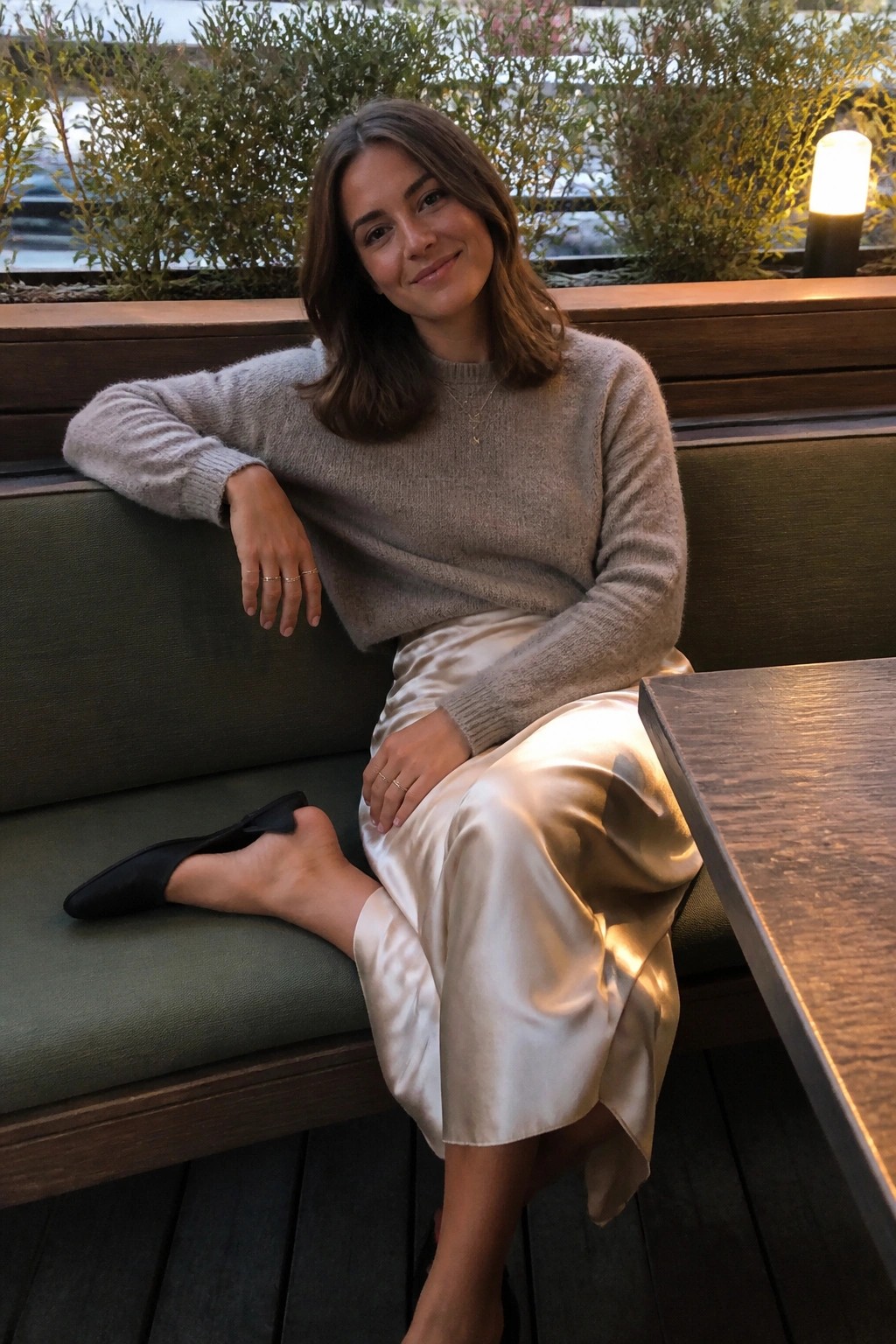 Woman in beige sweater and satin skirt seated on green bench smiling
