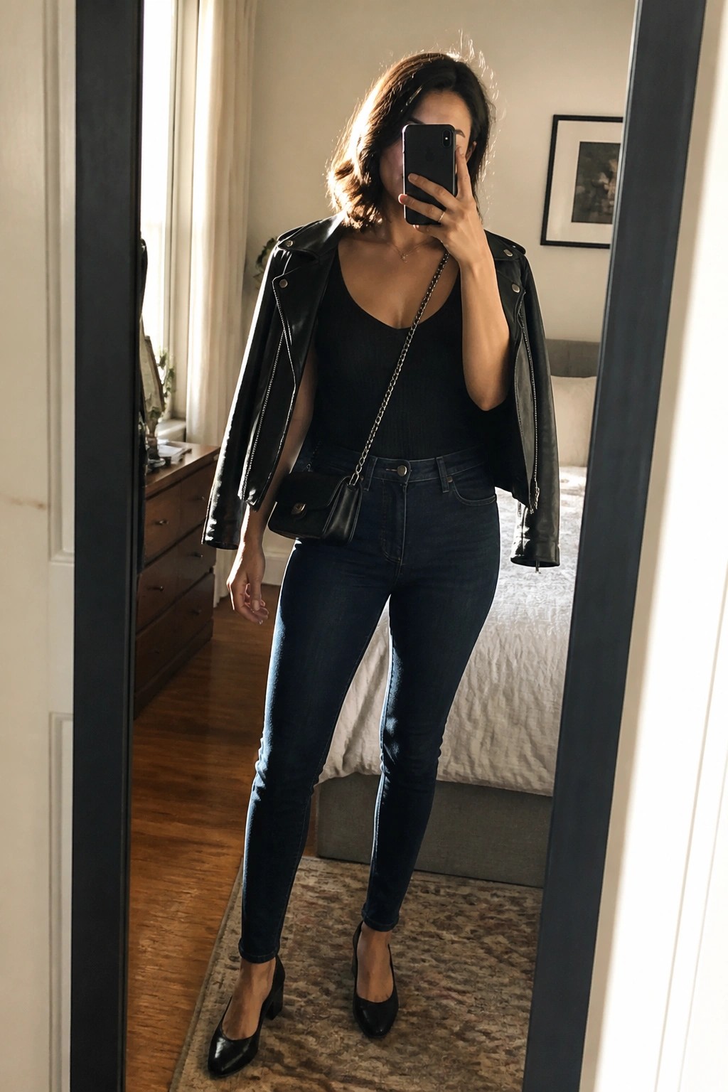 Woman in black leather jacket and jeans taking bedroom mirror selfie with phone