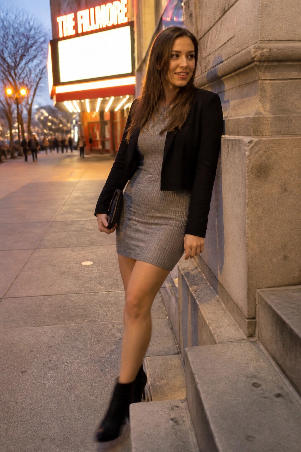 Woman wearing a short silver sequined dress under a black cropped blazer with black ankle boots and a clutch while posing on stone steps outside a theater marquee.