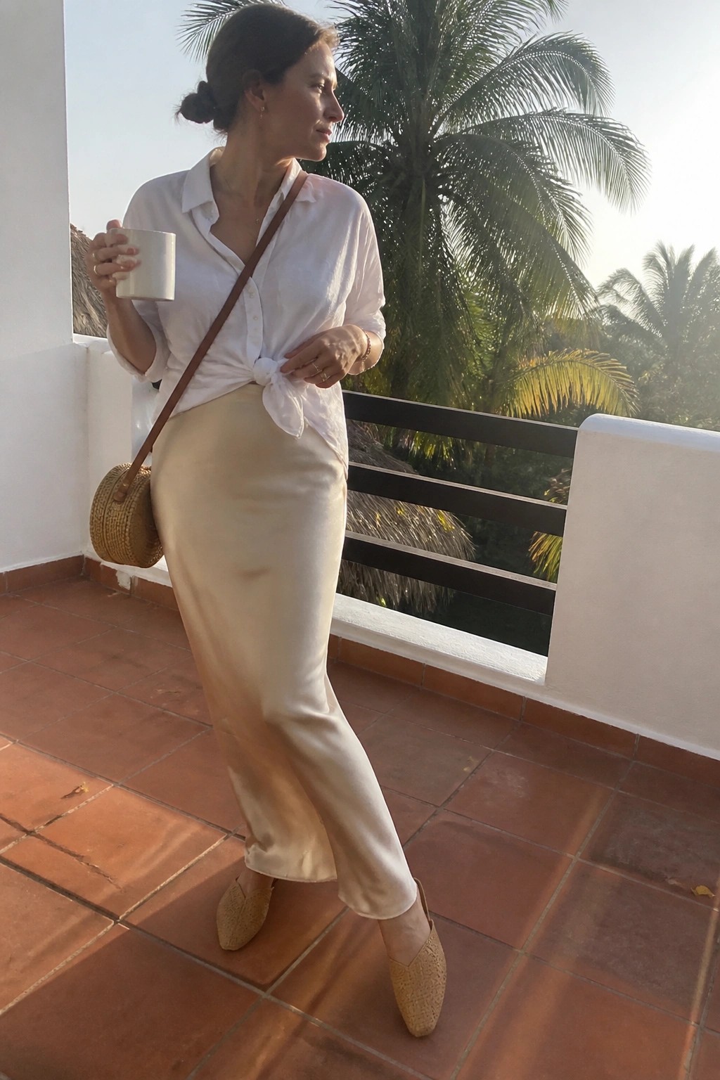 Woman in white tied shirt and beige skirt holding mug on tropical balcony