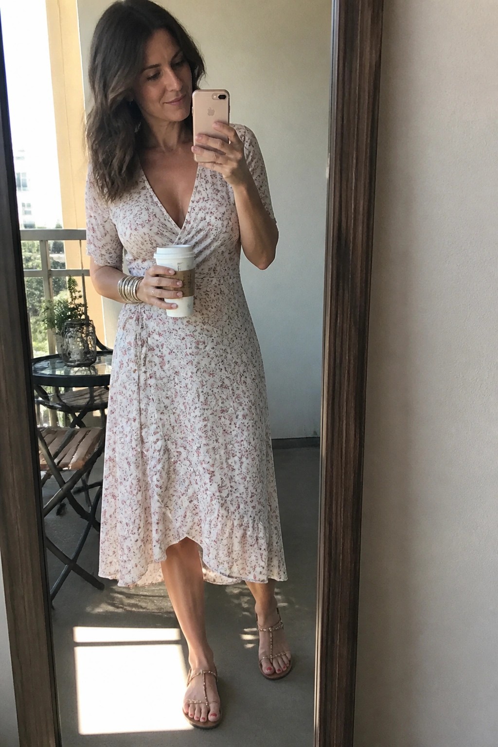 Woman in floral wrap dress taking mirror selfie while holding coffee on balcony