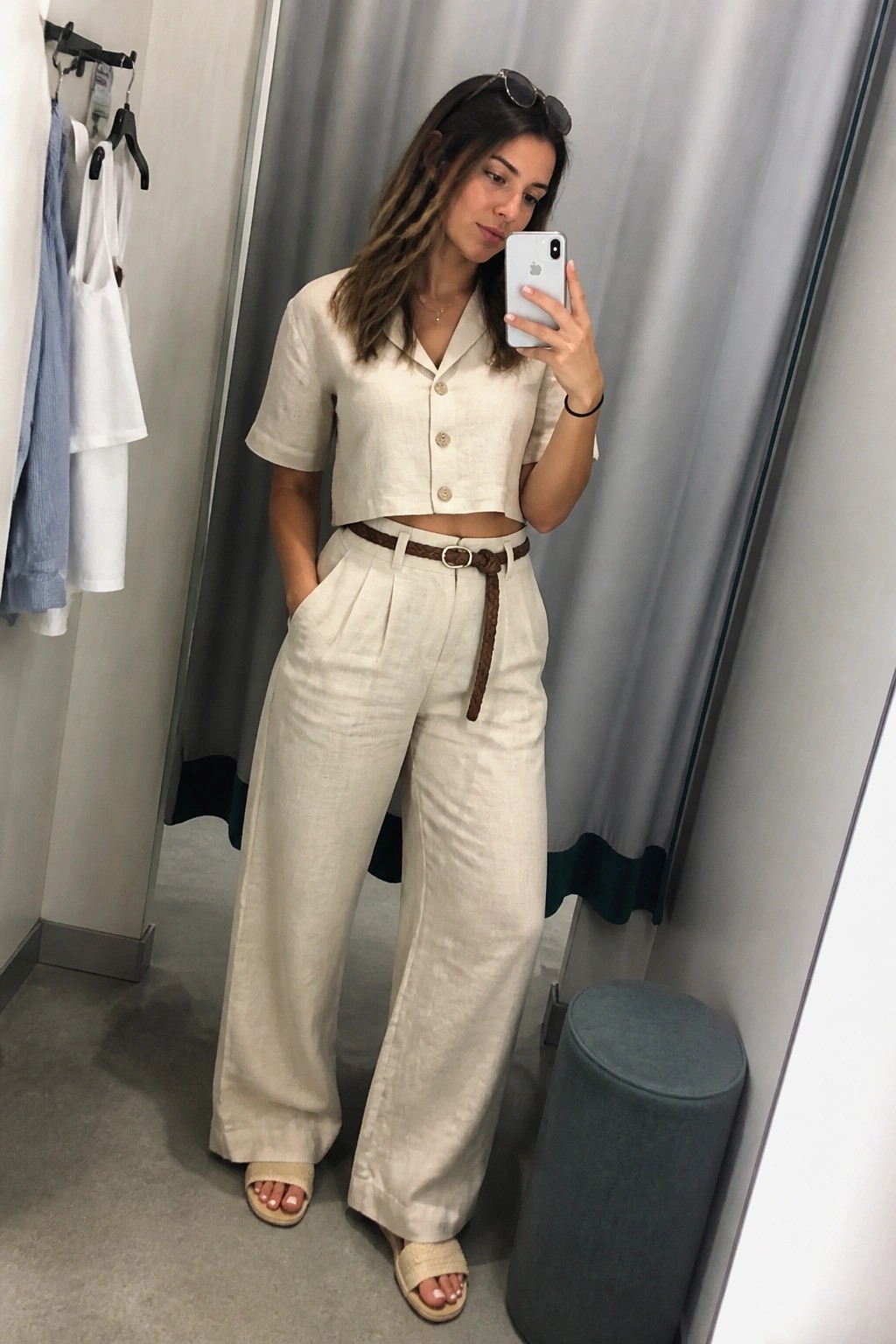 Woman wearing a cream linen cropped button-up shirt and matching wide-leg pants with a brown braided belt, beige woven slides, and sunglasses on her head while taking a mirror selfie in a fitting room.