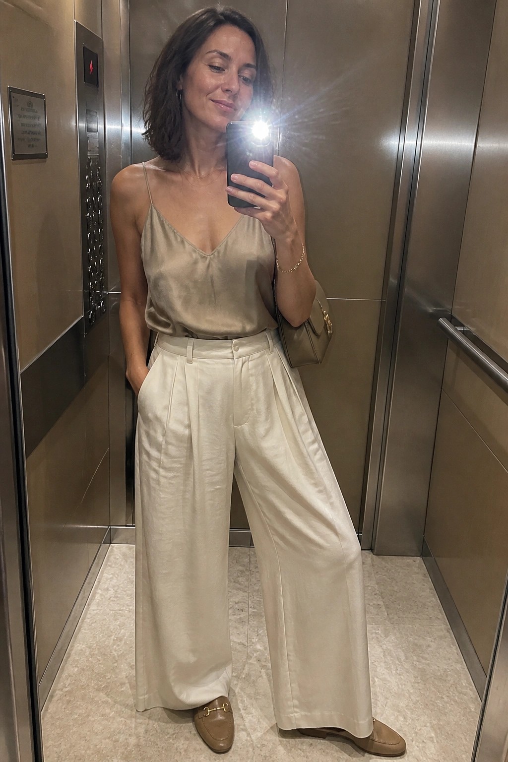 Woman in beige satin top and white wide-leg pants taking elevator selfie