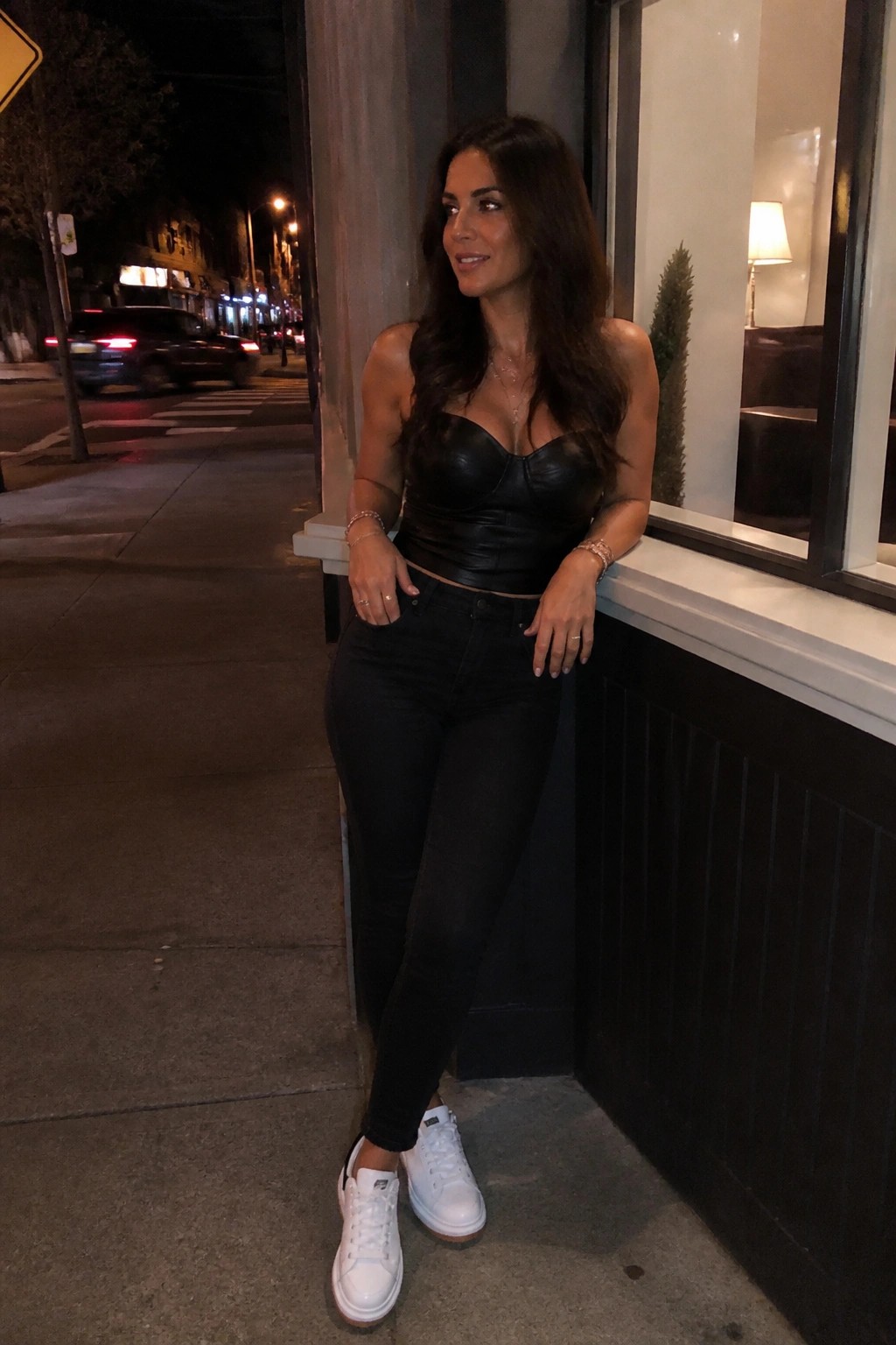 Woman in black leather crop top and jeans leaning on city window at night