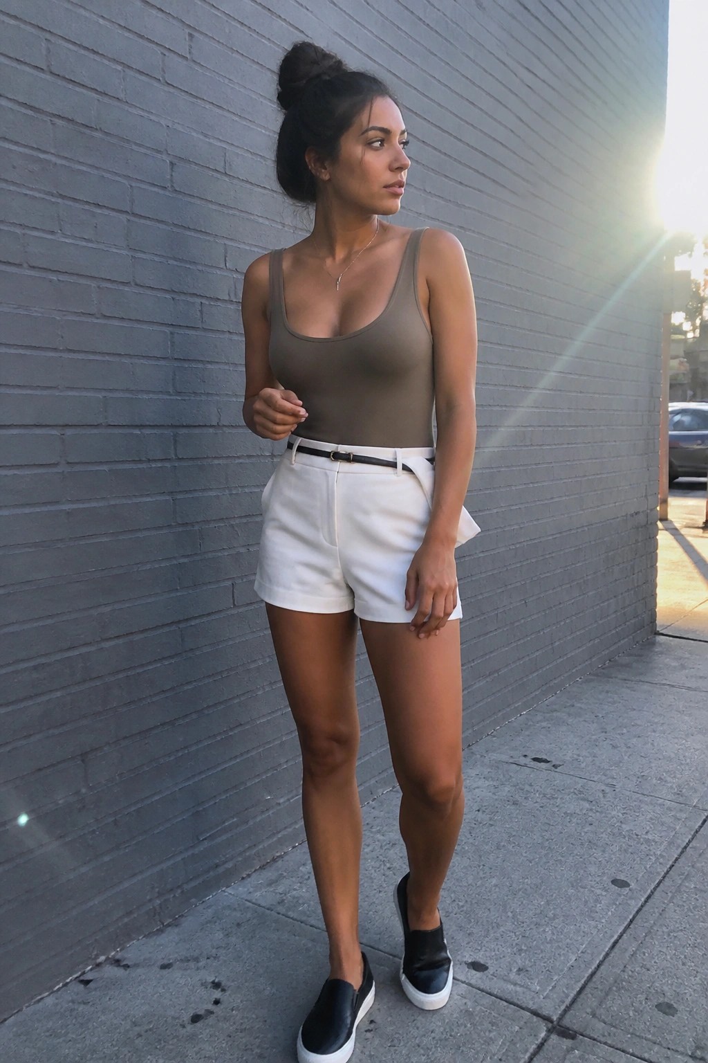 Woman in taupe sleeveless bodysuit tucked into white shorts with black belt and black slip-on sneakers standing against gray brick wall